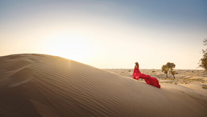 Fototapeta na wymiar Desert adventure. Young arabian Woman in red silk dress in sands dunes of UAE desert at sunset, fantastic view. The Dubai Desert Conservation Reserve, United Arab Emirates.