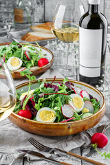 Summer fresh salad with eggs, radish, cucumbers and greens, arugula, leaves of lettuce in a plate on light wooden background served with glasses, wine, flowers. Healthy vegan food, clean eating