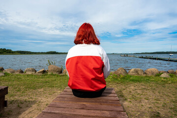 girl with red hair sits with her back to the camera on the shore of the lake