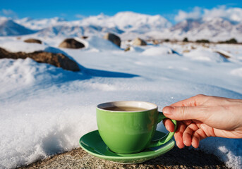 Woman hand holds green teacup on mountains background in winter - obrazy, fototapety, plakaty