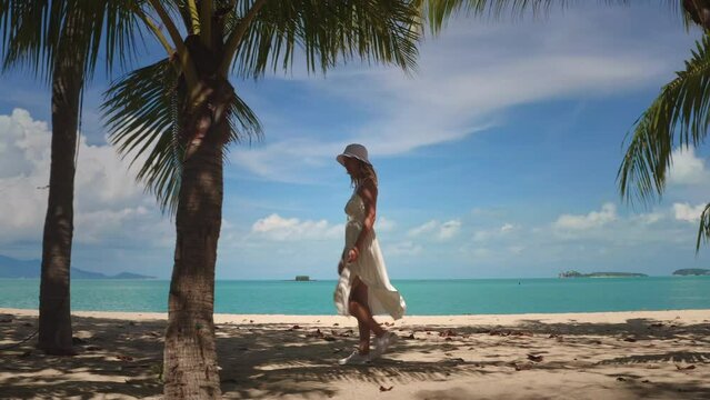 Elegant female in white dress walking tropical beach under palm trees, turquoise water, blue sky. Tourist woman relax and enjoy outdoor lifestyle travel on summer holiday vacation. Thailand, Samui
