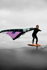 Male surfer balances himself while standing on a board with one hand holding the wings for wingfoiling