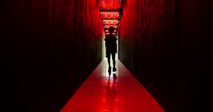 Silhouetted young boy moves towards us in slow motion down dark corridor bathed in dramatic red light. His unsettling movements add to eerie atmosphere of scene, creating sense of mystery and suspense