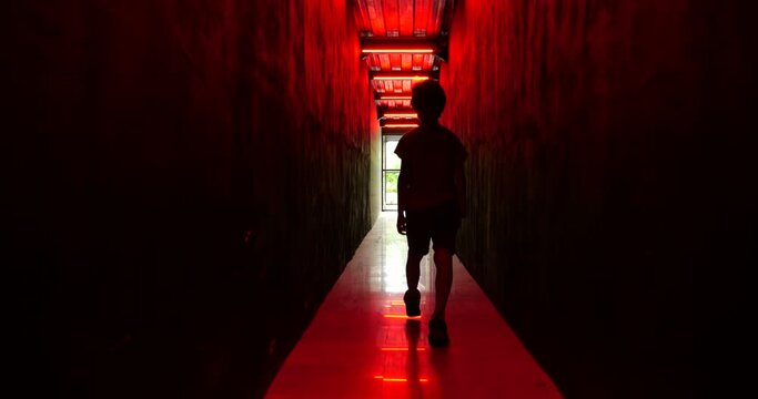 In dark corridor with black walls and red light, young boy walks away in slow motion. Camera follows him, capturing his silhouette in middle of passageway. Atmosphere is suspenseful and eerie