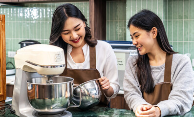 Two young asian women friends and aprons making cream for cupcakes, Using electric mixer, Preparing...