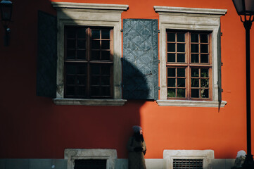 A young girl in a green down jacket and angora hat stands in profile near a terracotta-colored wall with windows and shutters in the rays of the setting sun with a dark shadow.Lviv, Armenian courtyard - obrazy, fototapety, plakaty