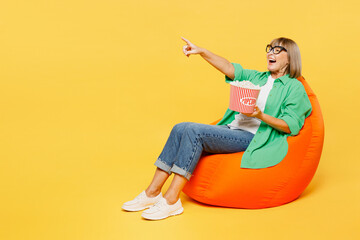 Full body surprised fun elderly woman 50s years old wear green shirt casual clothes sit in bag chair eat popcorn watch movie film point index finger aside on area isolated on plain yellow background.