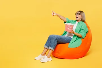 Poster Full body surprised fun elderly woman 50s years old wear green shirt casual clothes sit in bag chair eat popcorn watch movie film point index finger aside on area isolated on plain yellow background. © ViDi Studio
