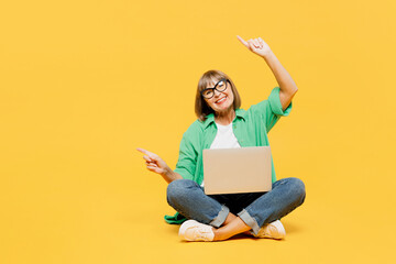 Full body elderly woman 50s years old wears green shirt glasses casual clothes sitting hold use work on laptop pc computer point aside on area isolated on plain yellow background. Lifestyle concept. - obrazy, fototapety, plakaty