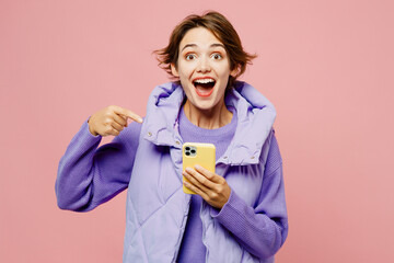 Young excited woman she wear purple vest sweatshirt casual clothes hold in hand use point finger on mobile cell phone isolated on plain pastel light pink background studio portrait. Lifestyle concept.