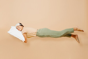 Full body sideways young calm Latin woman wears pyjamas jam sleep eye mask rest relax at home fly fall hover over air on pillow isolated on plain pastel beige background. Good mood night nap concept.