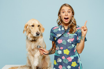 Young smart veterinarian woman she wear uniform heal exam retriever dog use stethoscope point index finger up isolated on plain pastel light blue background studio portrait. Pet health care concept.