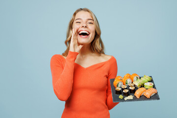 Young woman wears orange casual clothes scream sharing hot news about sales with hands near mouth hold eat raw fresh sushi roll served on black plate Japanese food isolated on plain blue background.