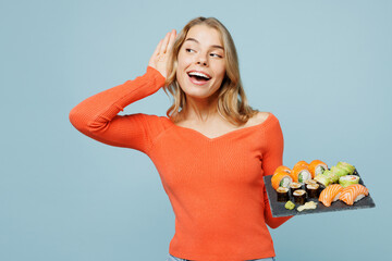 Young woman wear orange casual clothes try to hear you overhear listening intently hold eat raw fresh sushi roll served on black plate Japanese food isolated on plain blue background studio portrait.