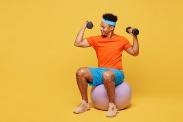 Full body happy young fitness trainer sporty man sportsman wearing orange t-shirt sit on fit ball hold dumbbells spend time in home gym isolated on plain yellow background. Workout sport abs concept.