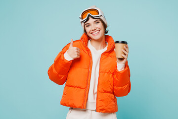 Skier young woman wears padded windbreaker jacket hat ski goggles mask hold takeaway delivery cup coffee to go travel rest spend weekend winter season in mountains isolated on plain blue background.
