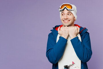 Obraz premium Amazed smiling happy man wear warm blue windbreaker jacket ski goggles mask hat hold snowboard look aside on area spend extreme weekend winter season in mountains isolated on plain purple background.