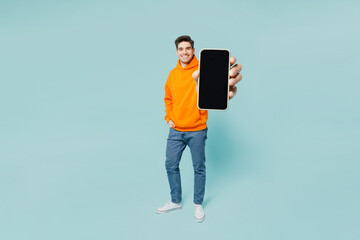 Full body young man he wears orange hoody casual clothes hold in hand use blank screen mobile cell phone isolated on plain pastel light blue cyan color background studio portrait. Lifestyle concept. - obrazy, fototapety, plakaty