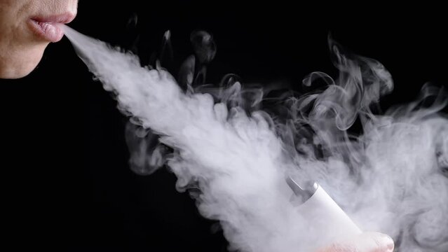 Girl Smoking a Vape or an E-Cigarette in a Dark Room on a Black Background. Close up. Blowing thick clouds of white smoke from the mouth. Texture. Filling. Smoking. Bad habits. Nicotine. Body part.