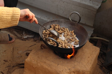An Indian Lady roasting ground nuts, peanuts or moong fali and salt in a black kadhai or pan on a clay chulha or desi stove outside, holding spoon
