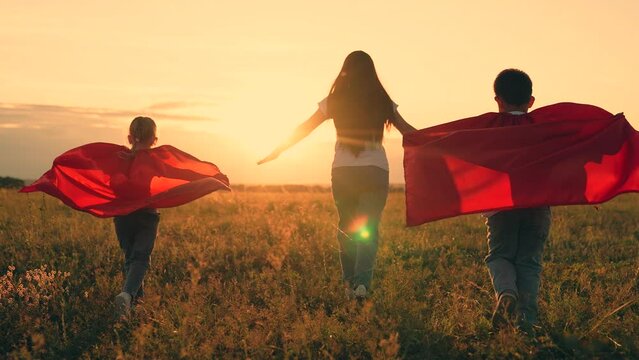 Mom, Super kids dream of becoming superhero, flying in red cape. Mother daughter, son play superheroes in park. Mom child run in red raincoats, have fun outdoor. Family Carnival in sun, Halloween, sky