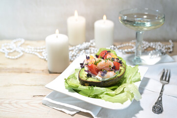 Salad from shrimps, tomatoes and mayonnaise filled in a halved avocado on a light rustic wooden table, wine glass, white candles and festive decoration, holiday meal, copy space
