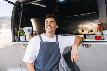 Cheerful food truck owner leaning counter. Young guy working as a chef in a food truck standing with closed eyes and smiling.
