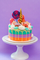 Green, purple and orange unicorn cake with mascarpone frosting roses, macaroons and golden horn. Copy space to side, plain purple background
