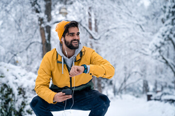Fototapeta na wymiar Confident handsome man with yellow beanie and windproof jacket tracking fitness progress on smart watch and using mobile phone while resting in squatting position after jogging.