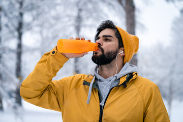 Exhausted handsome sportsman wearing yellow beanie and jacket drinking water from bottle after...