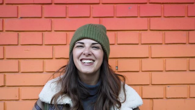 Young happy smiling woman smile while standing outdoor with wall background. POV happiness of student or freelancer female outside, wearing winter clothes wool hat and jacket looking at camera