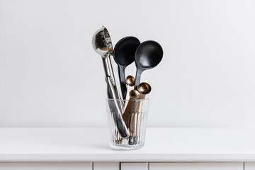 Assorted metal and black resin kitchen spoons and kitchen utensils inside a glass container on a white wooden cabinet