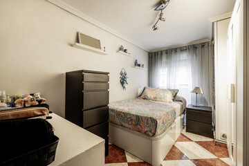 A bedroom with a single bed on a folding couch with very high drawers, furniture of various colors and stoneware floors