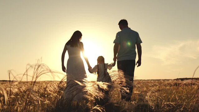 Silhouette of mother with father holding hands of funny daughter jumping over grass. Wife and husband walk looking at daughter strolling and jumping. Mother with father walking at sunset on field