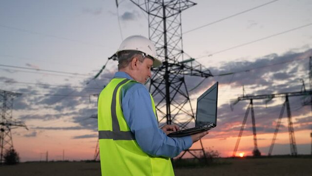 Man engineer uses laptop for power transmission lines condition diagnostic in evening. Electrician installs software for power distribution substation automation with laptop at twilight countryside