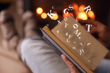 Man reading book with letters flying over it near fireplace at home, closeup