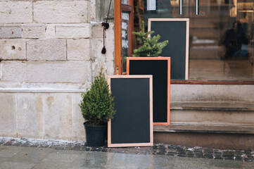 Old black wooden billboards stand on granite stones in the doorway near the Christmas tree. Outdoor advertising concept for cafes and shops. Copy space, mockup, layout.