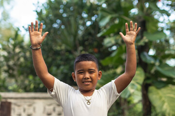 A carefree young asian boy raises his hands in the air in joy. Outdoor garden scene.