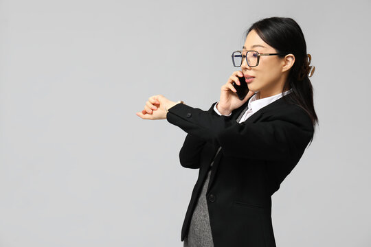 Portrait of young Asian businesswoman talking by phone and checking time on grey background
