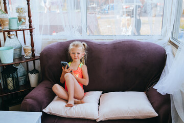 Curious young girl sitting on a sofa, holding a smartphone, by a window adorned with tulle curtains...