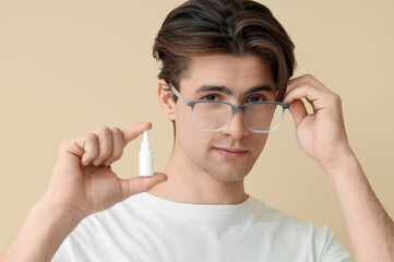 Young man in glasses with eye drops on beige background, closeup. Glaucoma awareness month