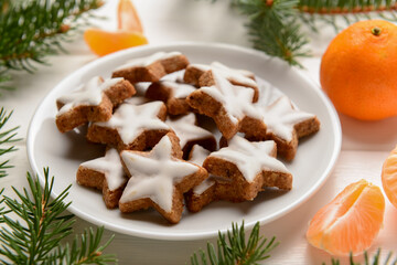 Plate with delicious stars shaped Christmas cookies, mandarins and fir tree branches on light wooden background, closeup