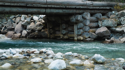 A strong wooden bridge made of logs and a clear mountain river. The water in the river is turquoise in colour. A small angle. The beauty of nature.