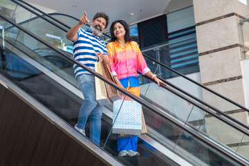 Happy indian couple holding on escalator handrail and riding escalator in shopping mall with paper bags, purchases in hands. Shopaholic concept.