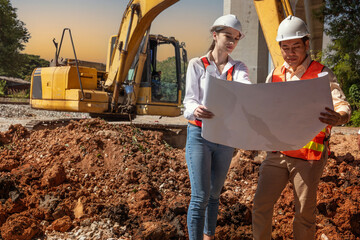 Asian male engineer and female worker wearing hard hats stand holding blueprints, talking and looking at construction drawings, behind a tractor digging dirt on a construction site. - Powered by Adobe