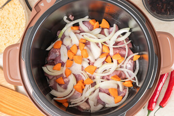 Frying meat with onions and carrots in a multicooker bowl. Modern, bright multi cooker with products. Cooking pilaf with meat in multicooker. Top view of multi cooker with vegetables and meat.