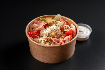 salad with chicken meat, bacon in paper take away container with sauce on black background. Healthy food concept, restaurant dish delivery.