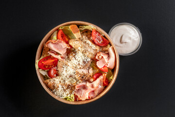 salad with chicken meat, bacon in paper take away container with sauce on black background. Healthy food concept, restaurant dish delivery.