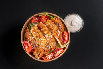 Snittel Caesar Salad in a paper takeaway container with sauce on a black background. Healthy eating concept, restaurant food delivery. View from above. Free space for your text.