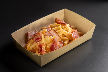 Fondue fried potatoes with cheese and crispy bacon on a black background, in a container for delivery. Deep fry potato or appetizer, fast food.